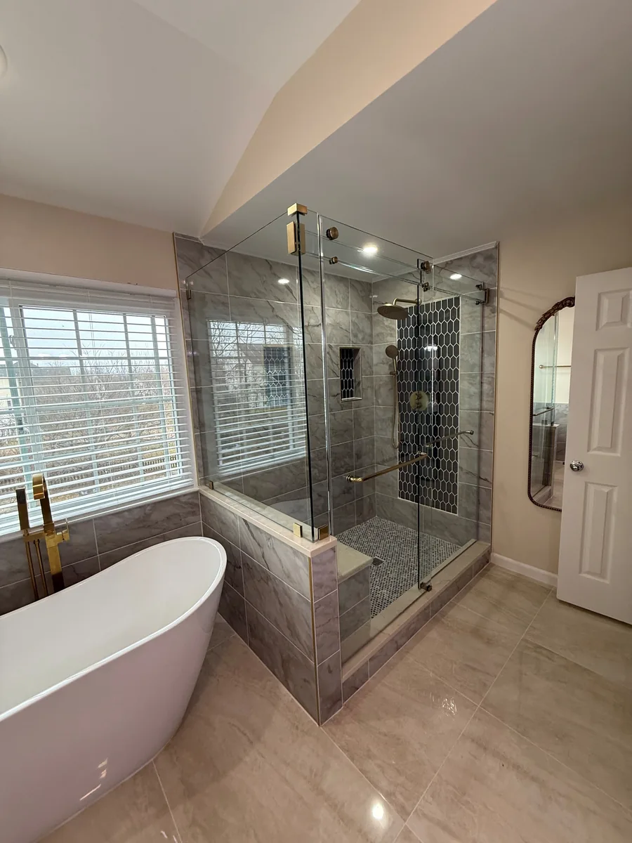 Corner glass shower with gold-tone sliding hardware and dark patterned tile beside a white freestanding bathtub.