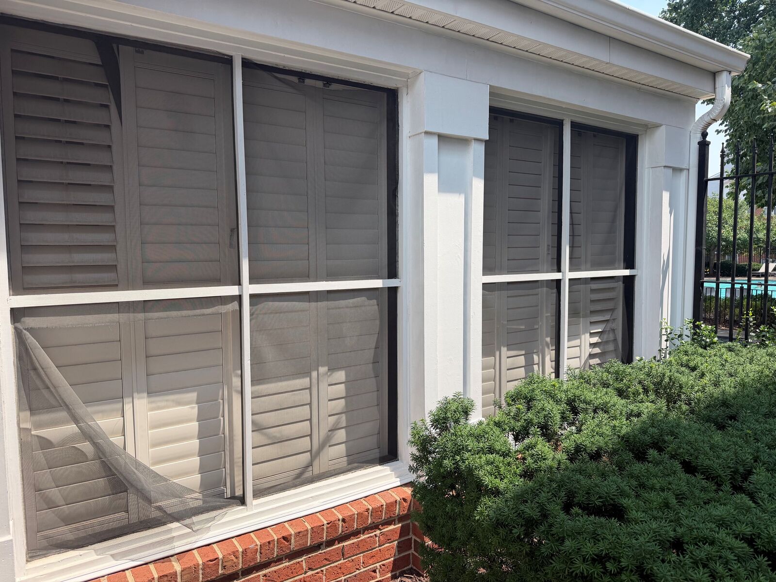 Torn porch screen hanging loose in front of white shutters above a brick base