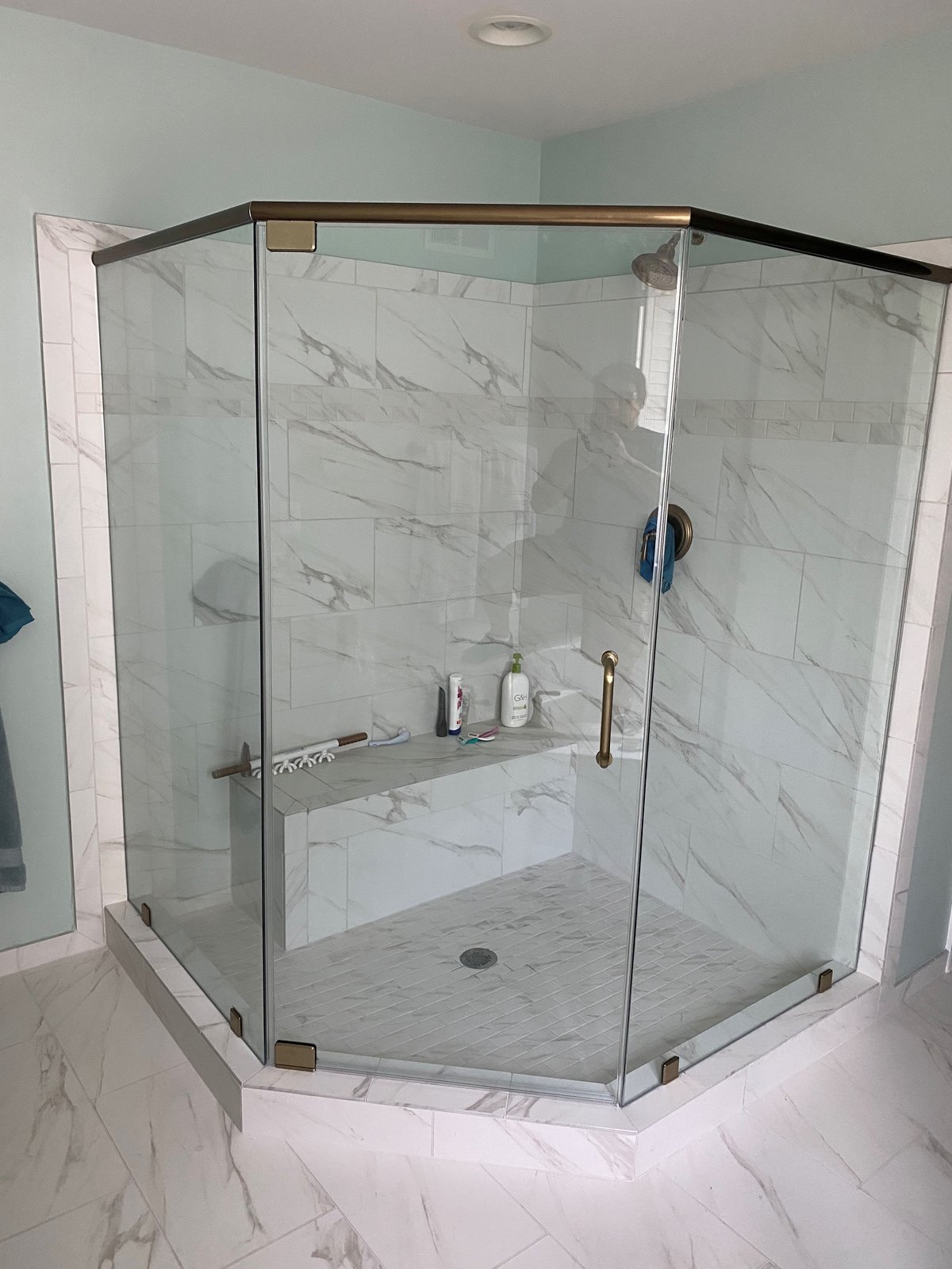 A neo-angle glass shower with a brass-tone header across the top and a tiled bench