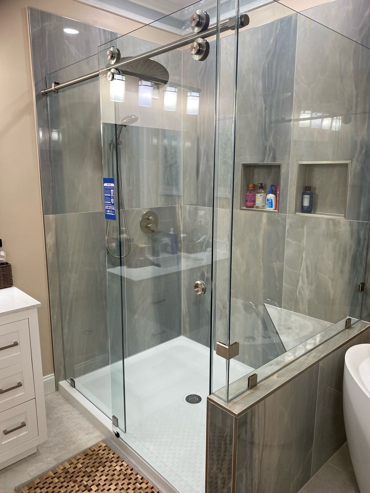 A frameless sliding shower door with an exposed roller track across the top, gray wall tile and a half wall